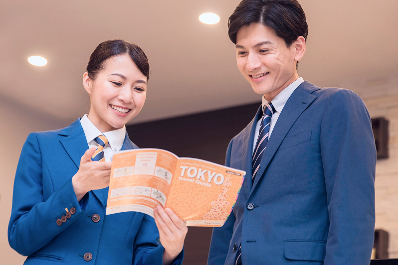 ホテルのフロントスタッフが笑顔で書類を手渡しながら、スーツ姿の男性と対話している。明るく清潔なロビーでの接客の様子。