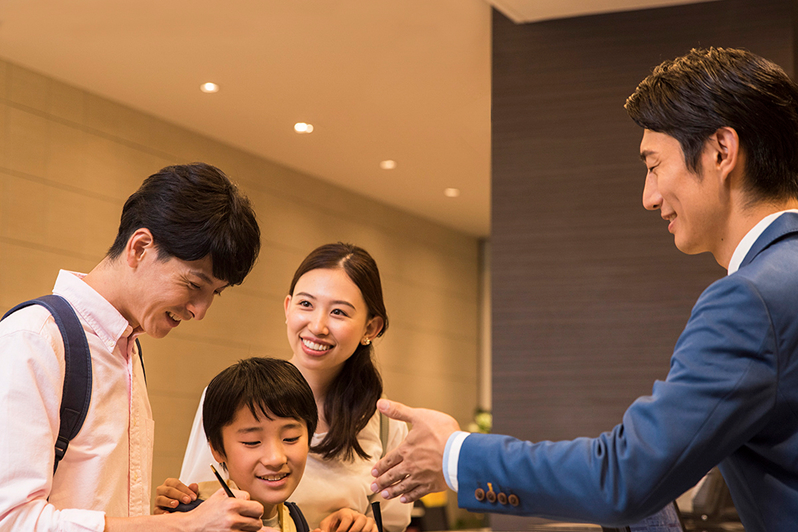 ホテルのフロント前で、笑顔のスタッフが上品に立ってお客様を迎えている。背後には『Comfort HOTEL』のロゴが見える。