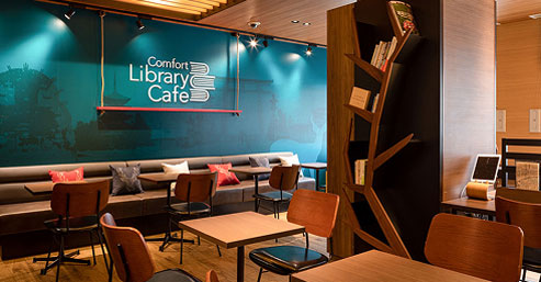 Comfort Library Cafe」のロゴが光る、落ち着いた雰囲気のカフェスペース。木製のテーブルと椅子が並び、奥にはソファ席も見える。