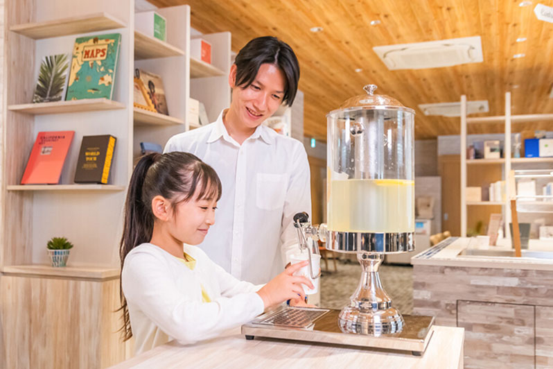 セルフサービスのドリンクコーナーで、男性が笑顔で女の子にレモンウォーターを注ぐ様子。木のインテリアが温かみを感じさせる。