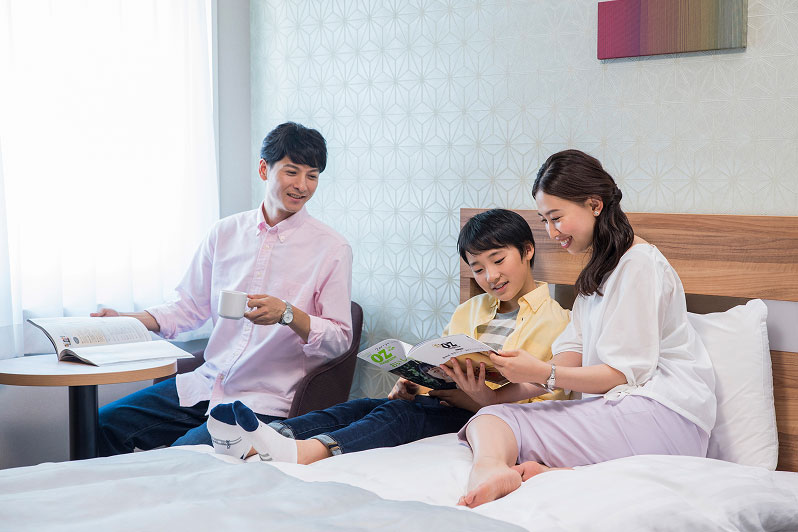 ホテルのベッドの上で、家族3人がリラックスしながら本を読んでいる。父親は雑誌を手に持ち、母親と娘は一緒に絵本を見ている。