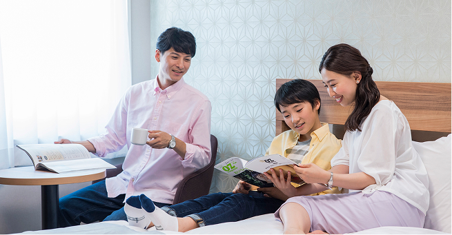 ベッドの上で母親と子どもがくつろぎながら一緒に本を読み、父親がベッド側の椅子に座って読書をしている様子。