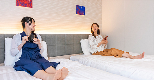 女性二人がツインベッドの上でそれぞれ本を読んだりドリンクを飲んだりしている。