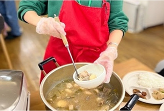 エプロンを着けた人がおたまで汁物をよそっている。食事提供の準備をしている様子。