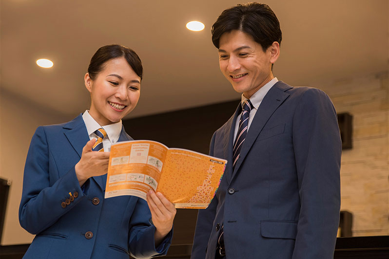 ホテルの受付係が青い制服と黄色いネクタイを着て、フロントデスクで電話をしながら微笑んでいます。背景には「Comfort Hotel」のロゴと、キャンドルが置かれた装飾的な棚が付いた白い壁があります。